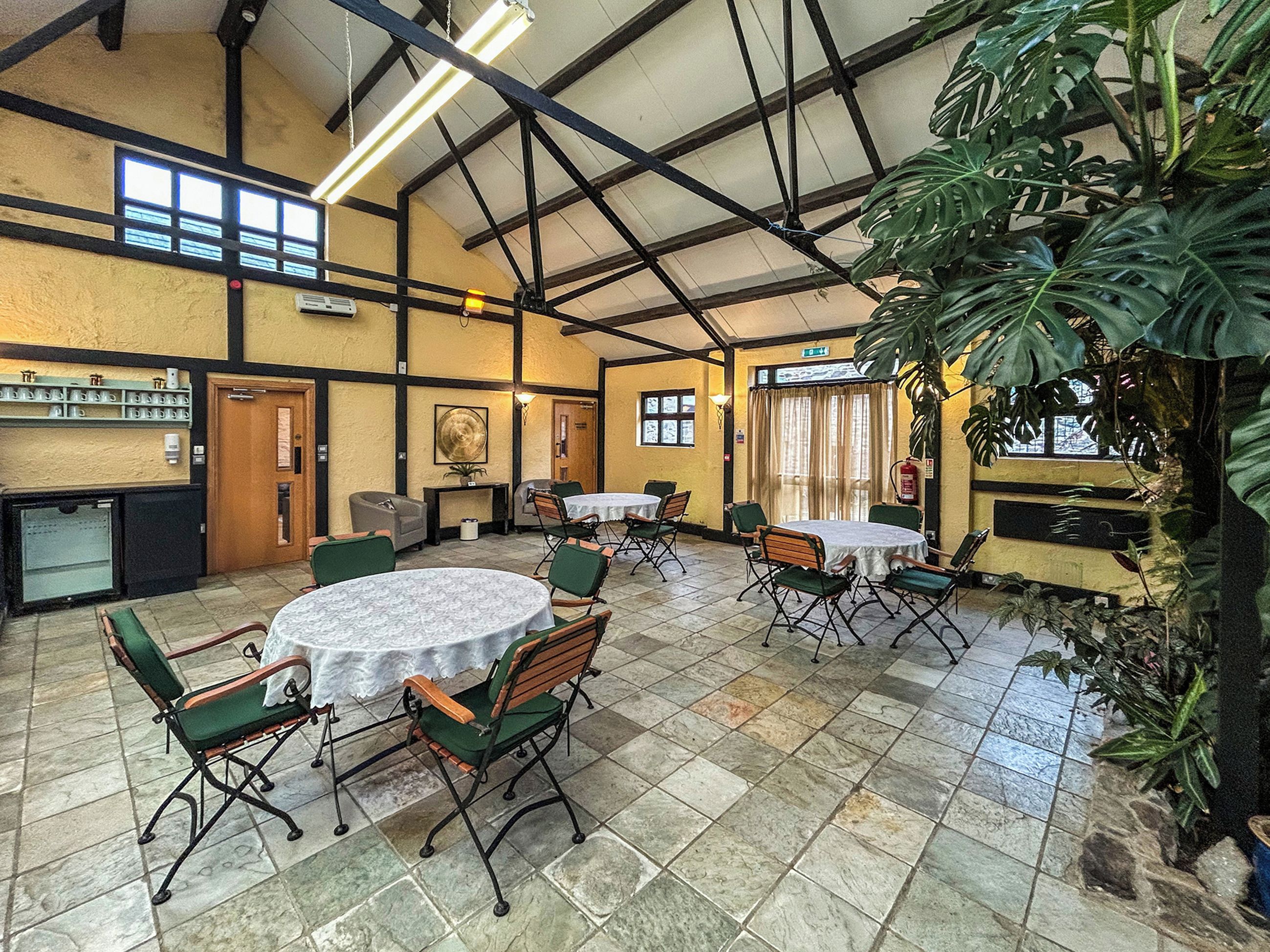 Un vaste espace de restauration intérieur doté de hauts plafonds, de nombreuses tables et chaises, ainsi que d’une grande plante et d’un extincteur.