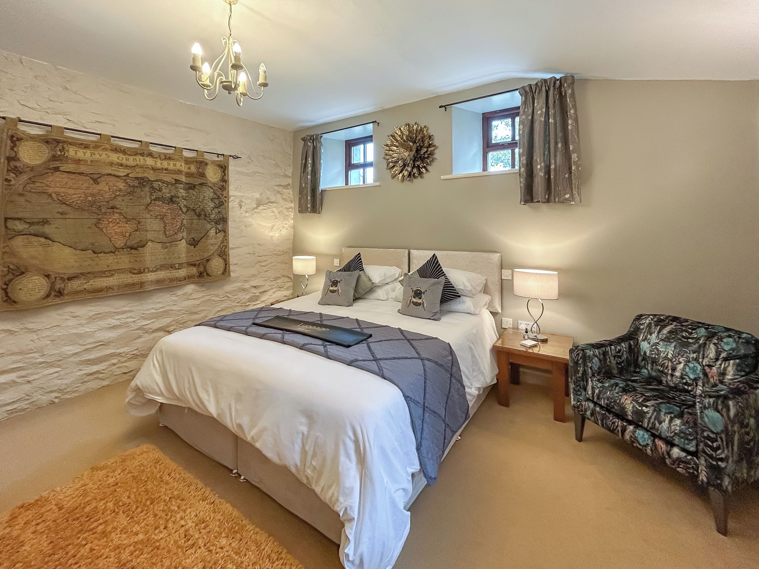 Une chambre à coucher avec un grand lit, une table de chevet, une chaise, un lustre, une carte accrochée au mur et une fenêtre avec des rideaux.