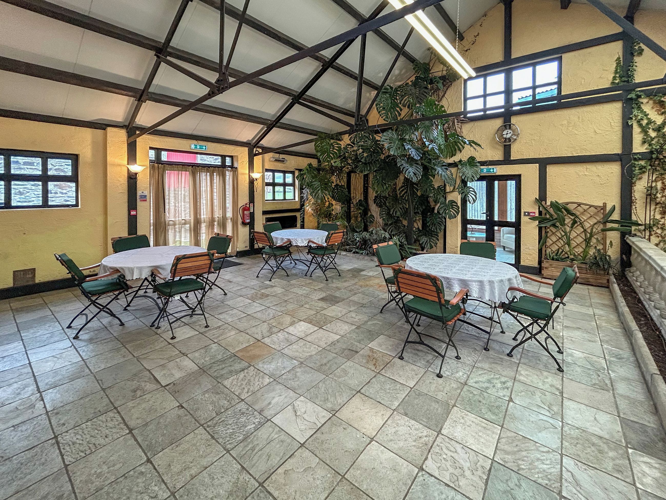 A spacious indoor area with multiple round tables and chairs, large windows, and potted plants.