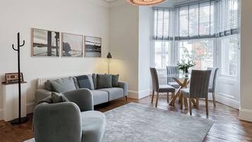 A living room with a grey sofa, a coffee table, and a dining area with chairs. There is a large window with blinds, a floor lamp, and wall art.