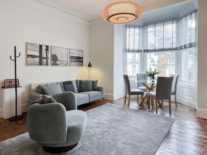 Een woonkamer met een grijze bank, een salontafel en een eethoek met stoelen. Er is een groot raam met zonwering, een vloerlamp en wanddecoratie.