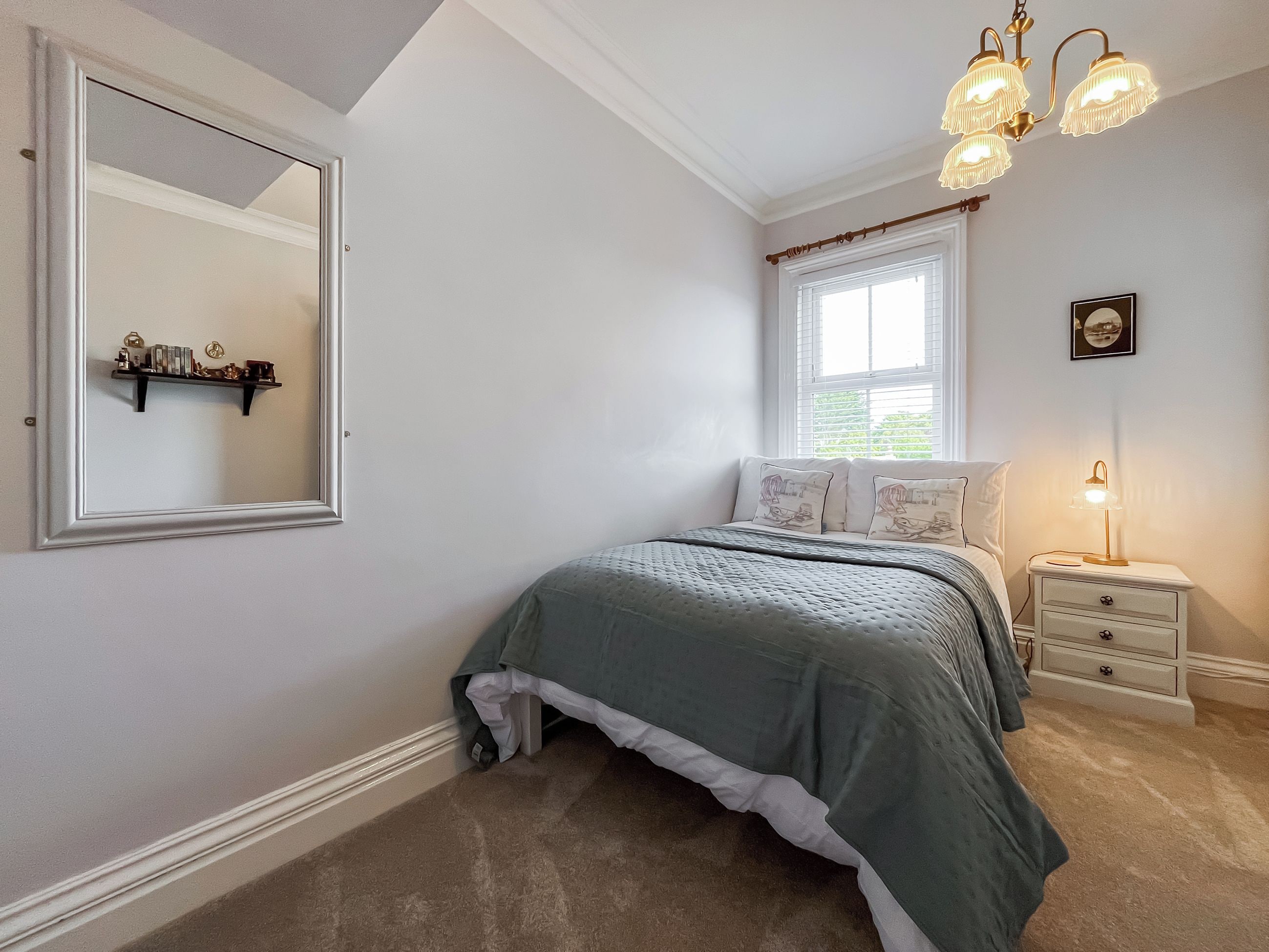 A bedroom with a bed, a nightstand, a lamp, a window with blinds, a mirror, and a shelf with decorative items.