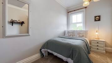 A bedroom with a bed, a nightstand, a lamp, a window with blinds, a mirror, and a shelf with decorative items.