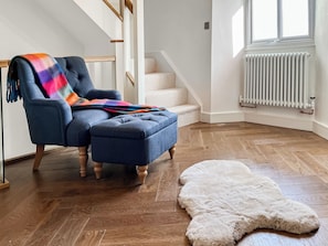 Une pièce avec un fauteuil bleu, un tapis blanc et moelleux, un plancher en bois, un radiateur blanc et un escalier.