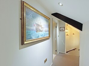 Um corredor com um quadro de um navio na parede, uma porta que dá para outro cômodo e uma escada com corrimão preto.