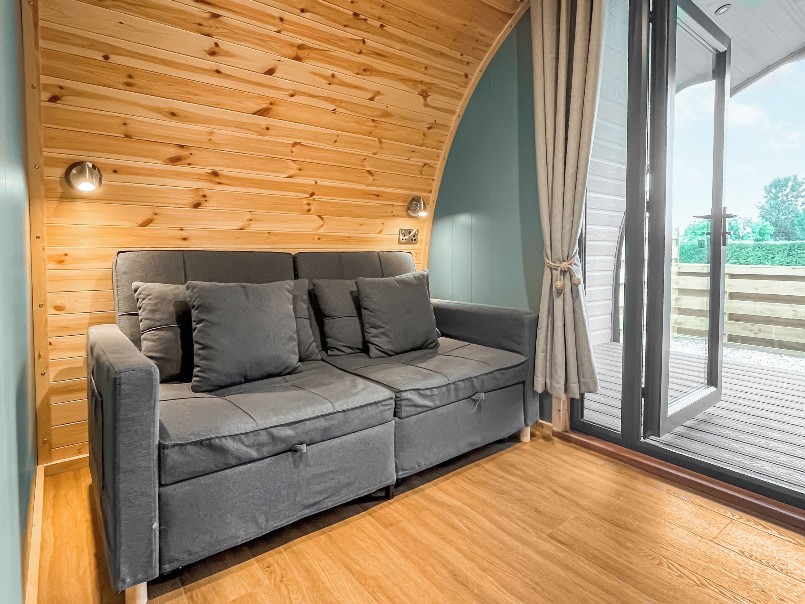 Ein graues Sofa mit Kissen vor einer Holzwand, ein Fenster mit Vorhängen und Blick auf Grünfläche draußen.