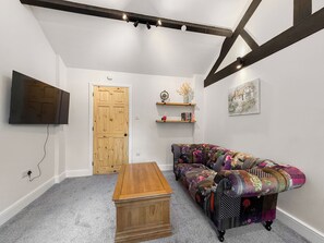 A room with a wooden coffee table, a colorful sofa, a flat-screen TV mounted on the wall, a wooden door, and a shelf with decorative items. - 1 bedroom accommodation in Oswestry (Oswestry)