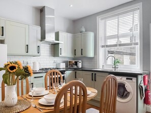 A modern kitchen with white cabinets, a dining table set for four, and a built-in washing machine. - Castle Street Cottage (Skipton)