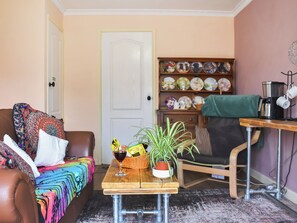 A cozy living room with a brown leather sofa, a colorful blanket, a wooden coffee table with a basket and a glass of red wine, a green potted plant, and a bookshelf with decorative plates. - We love the peaceful setting and sunny south-facing garden. (Tittleshall)