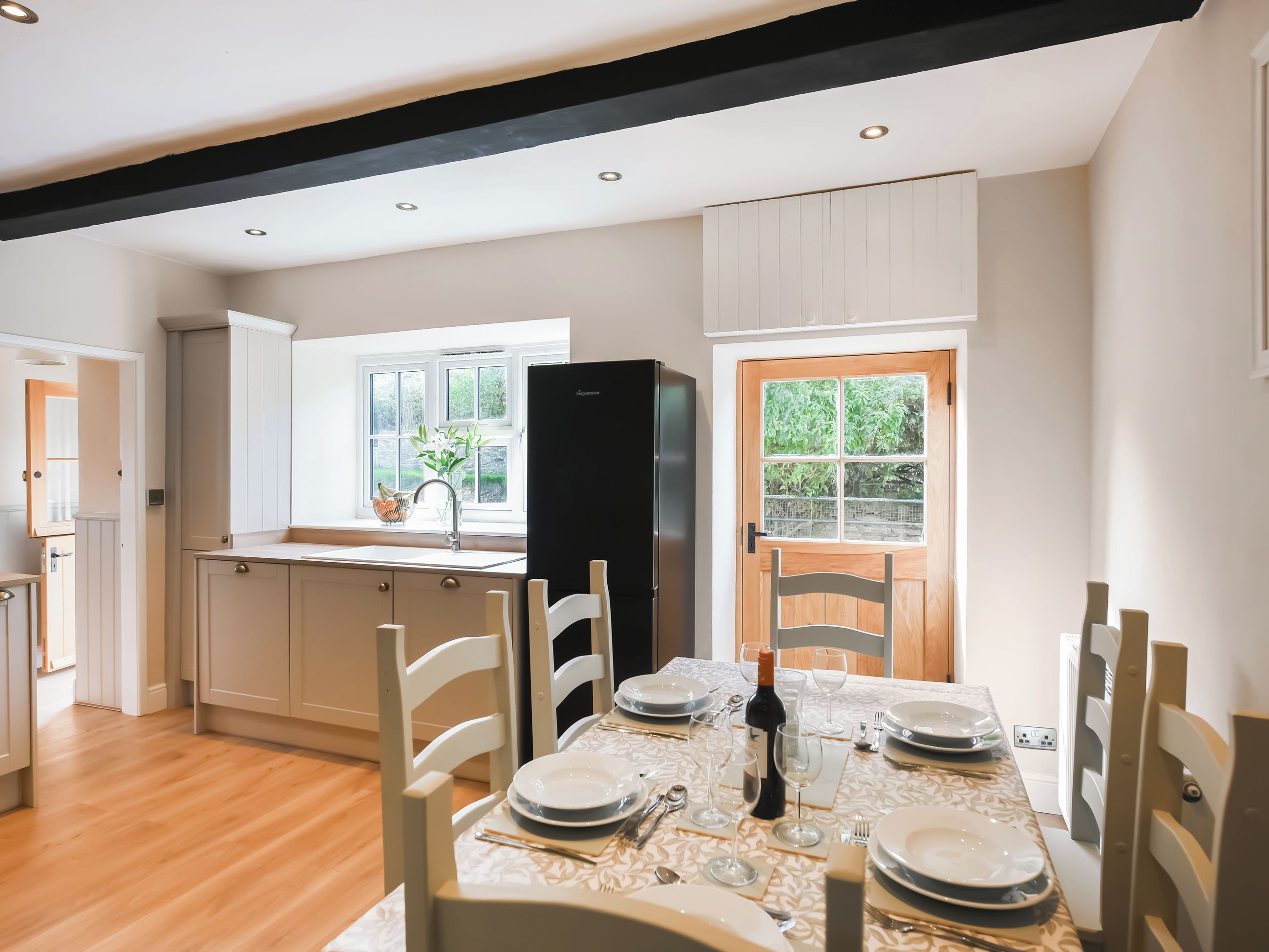 Une cuisine moderne avec un coin repas aménagé, comprenant des meubles en bois, un réfrigérateur noir et une fenêtre donnant sur un paysage verdoyant.