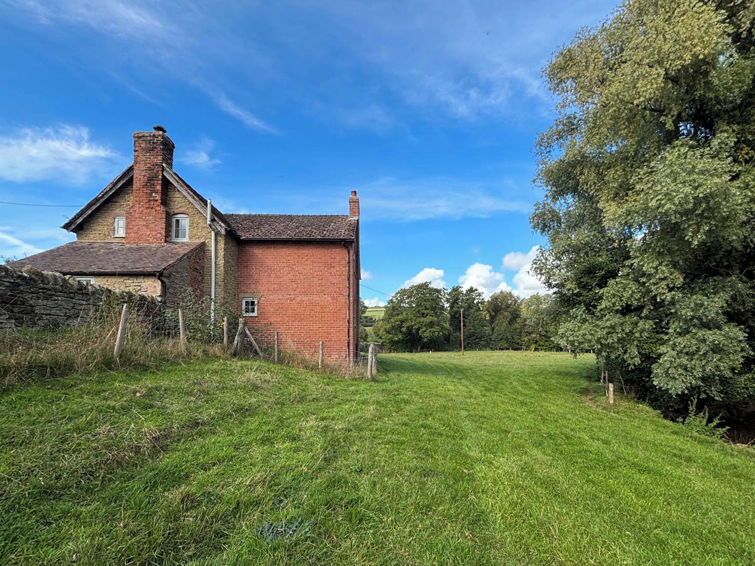 Une maison de campagne avec une façade en briques, un mur en pierre et une cour couverte d’herbe.