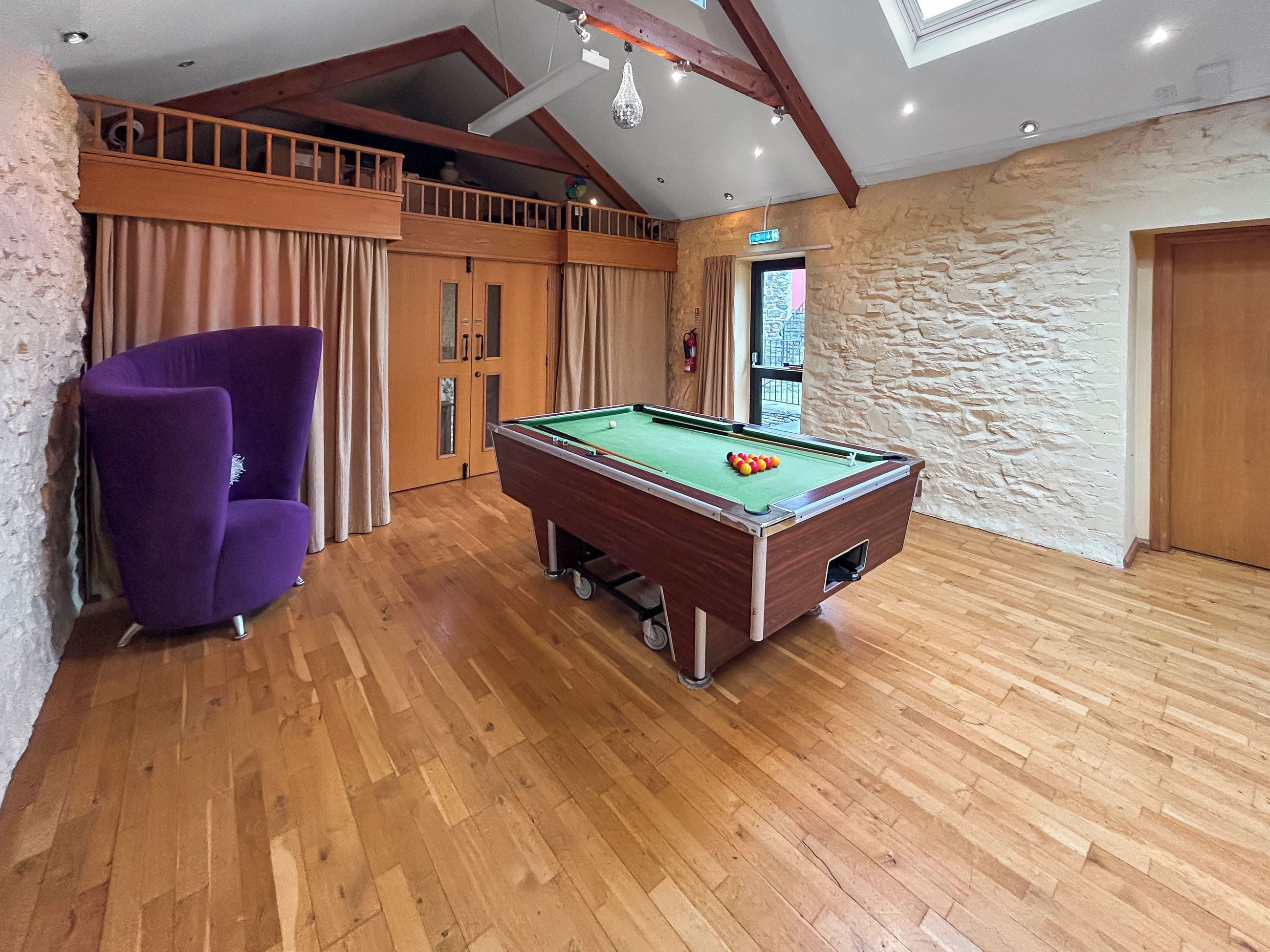 A room with a pool table, wooden flooring, a purple armchair, and a stone wall.