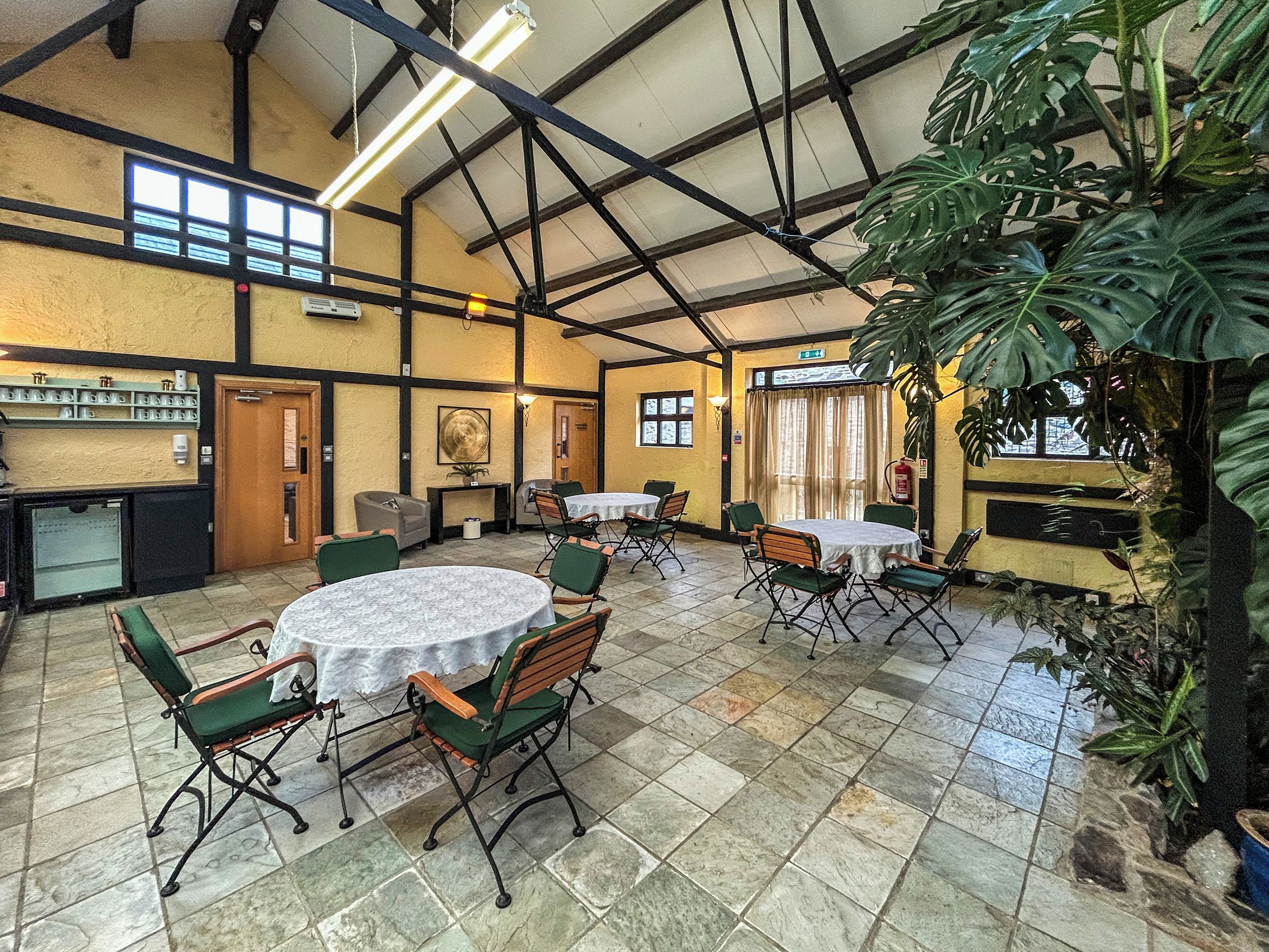 A spacious indoor area with multiple round tables and chairs, a bar area, and large windows allowing natural light.