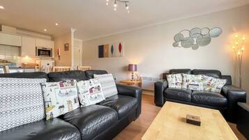 A modern living room with a black leather sofa, a wooden coffee table, and a dining area in the background.