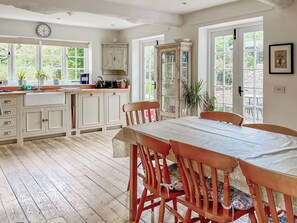 A kitchen with wooden cabinets, a dining table, chairs, a clock, and a window with a view of greenery. - Old Post Office Cottage (Weymouth)