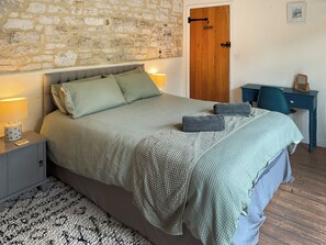 A bedroom with a stone wall, a wooden door, a bed with green bedding, a desk with a chair, and a rug on the floor. - Old Post Office Cottage (Weymouth)