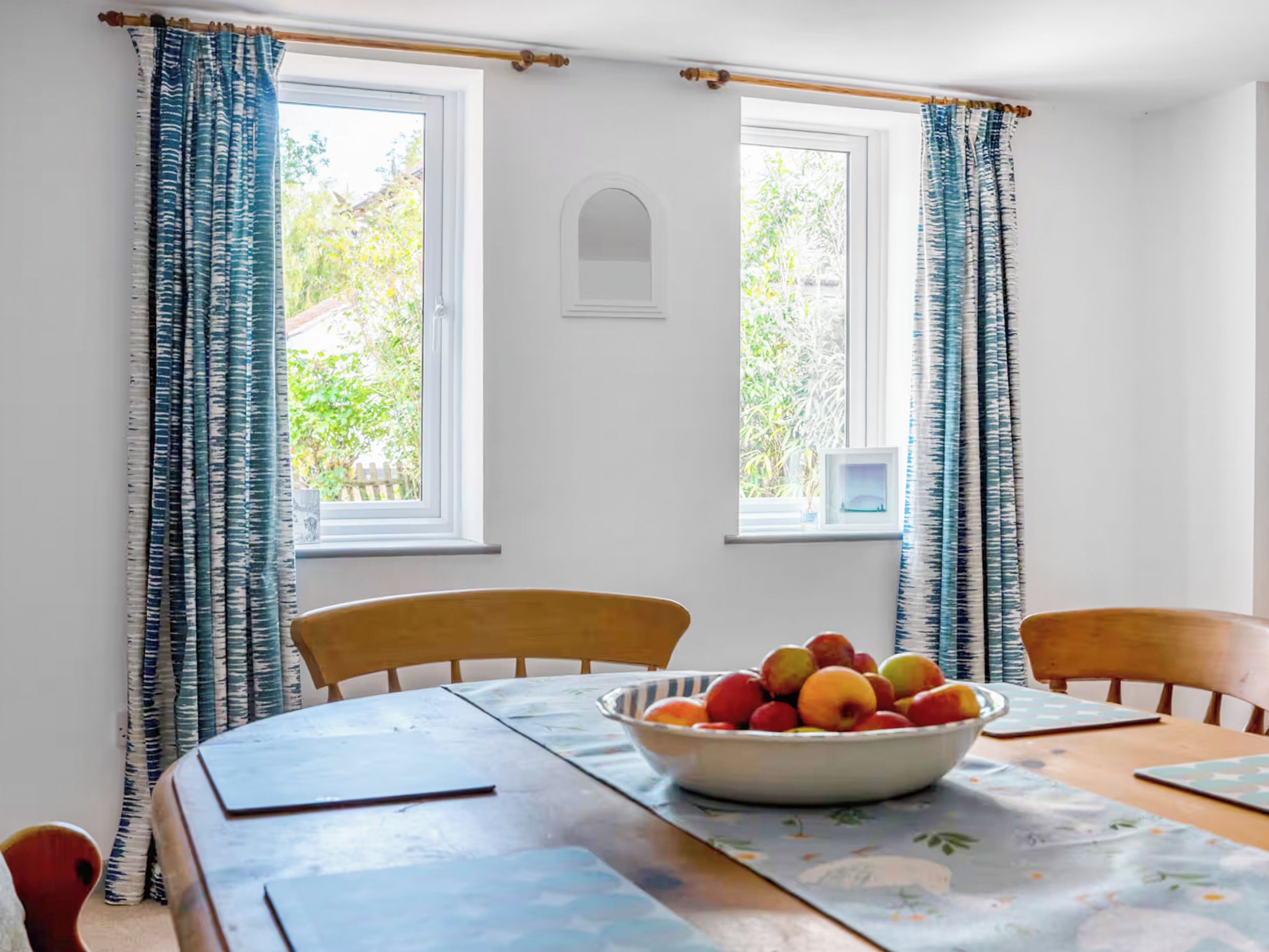 Un comedor con una mesa, sillas, un cuenco de fruta y dos ventanas con cortinas.