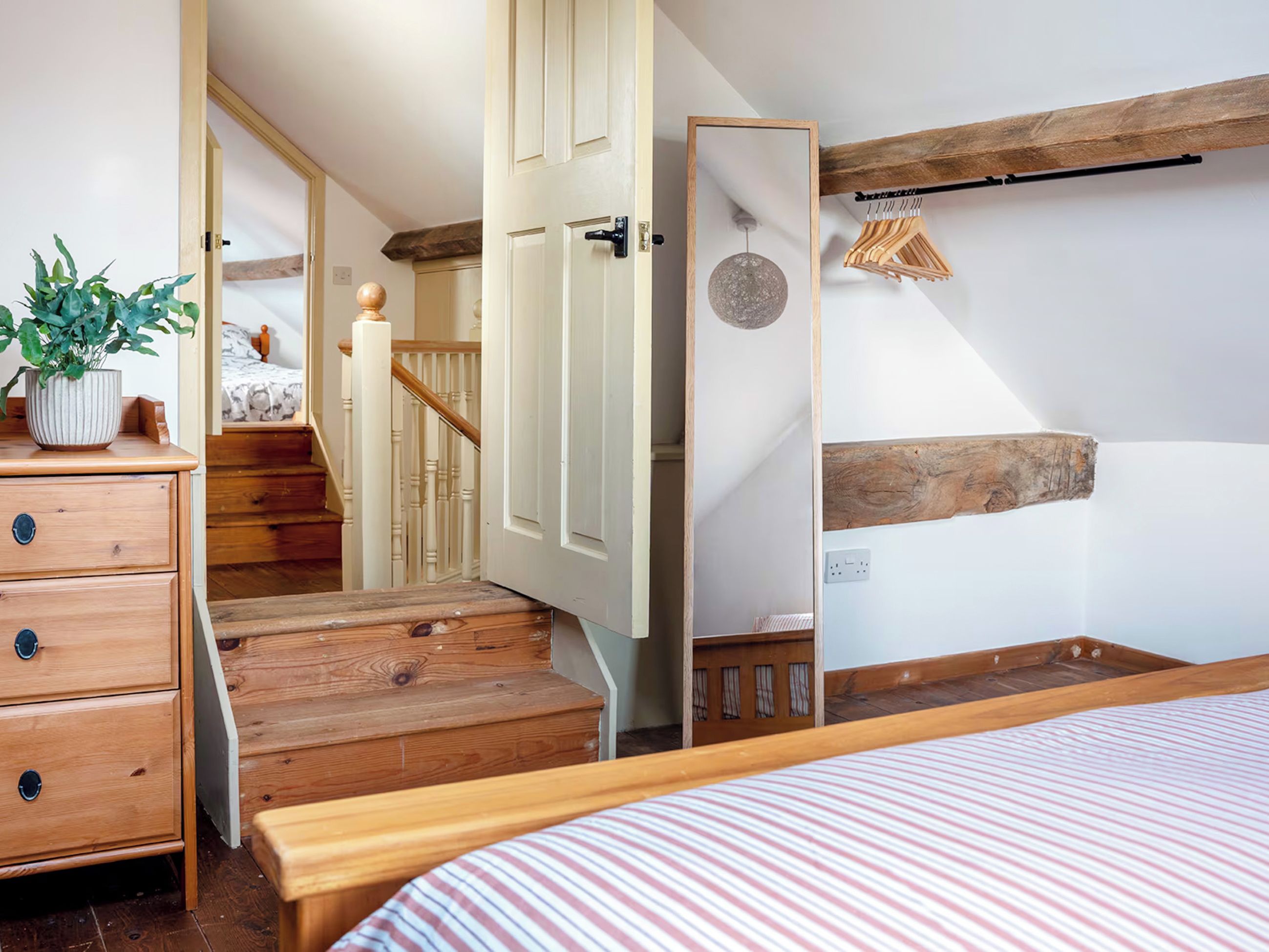 A bedroom with a wooden bed, a dresser with drawers, a potted plant, and a staircase leading to a bedroom.