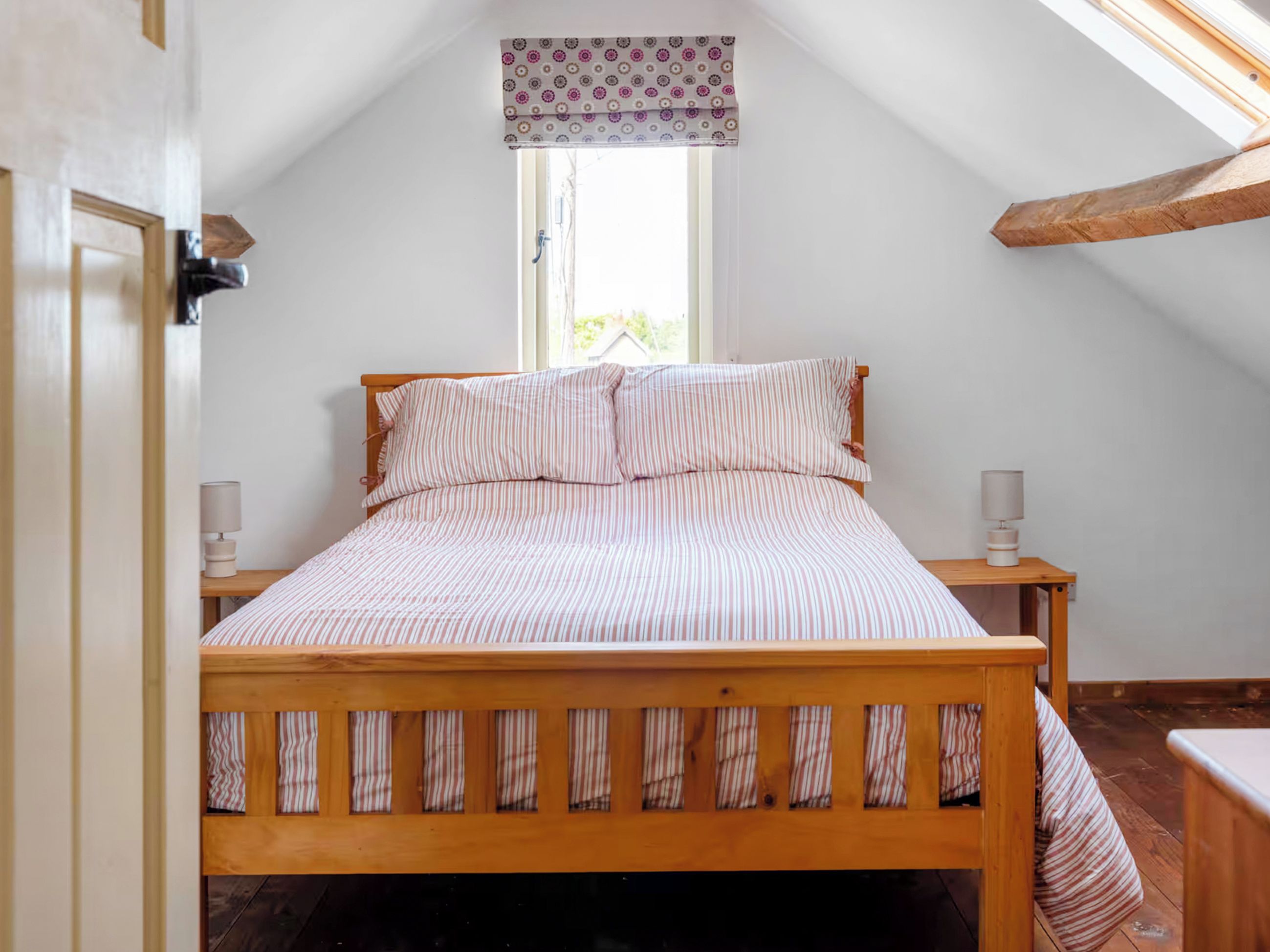A bedroom with a wooden bed, two bedside tables with lamps, a window with a patterned valance, and a sloped ceiling.