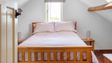 A bedroom with a wooden bed, two bedside tables with lamps, a window with a patterned valance, and a sloped ceiling.