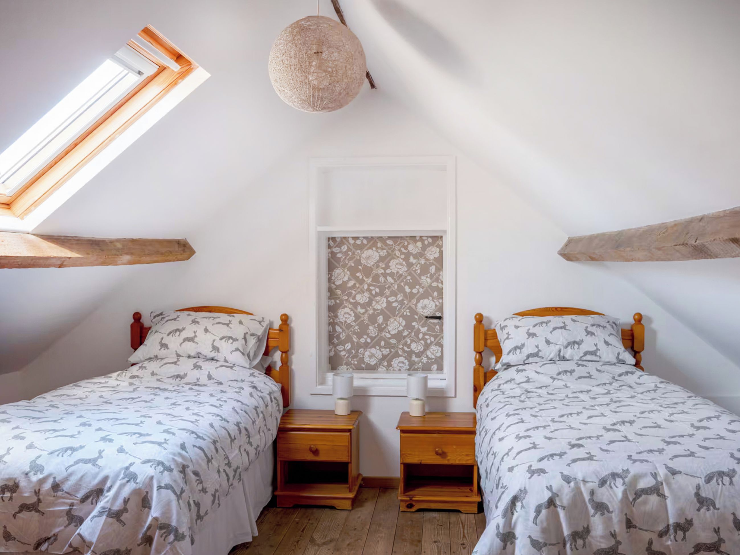A bedroom with two beds, wooden headboards, a window with floral wallpaper, and a hanging light fixture.