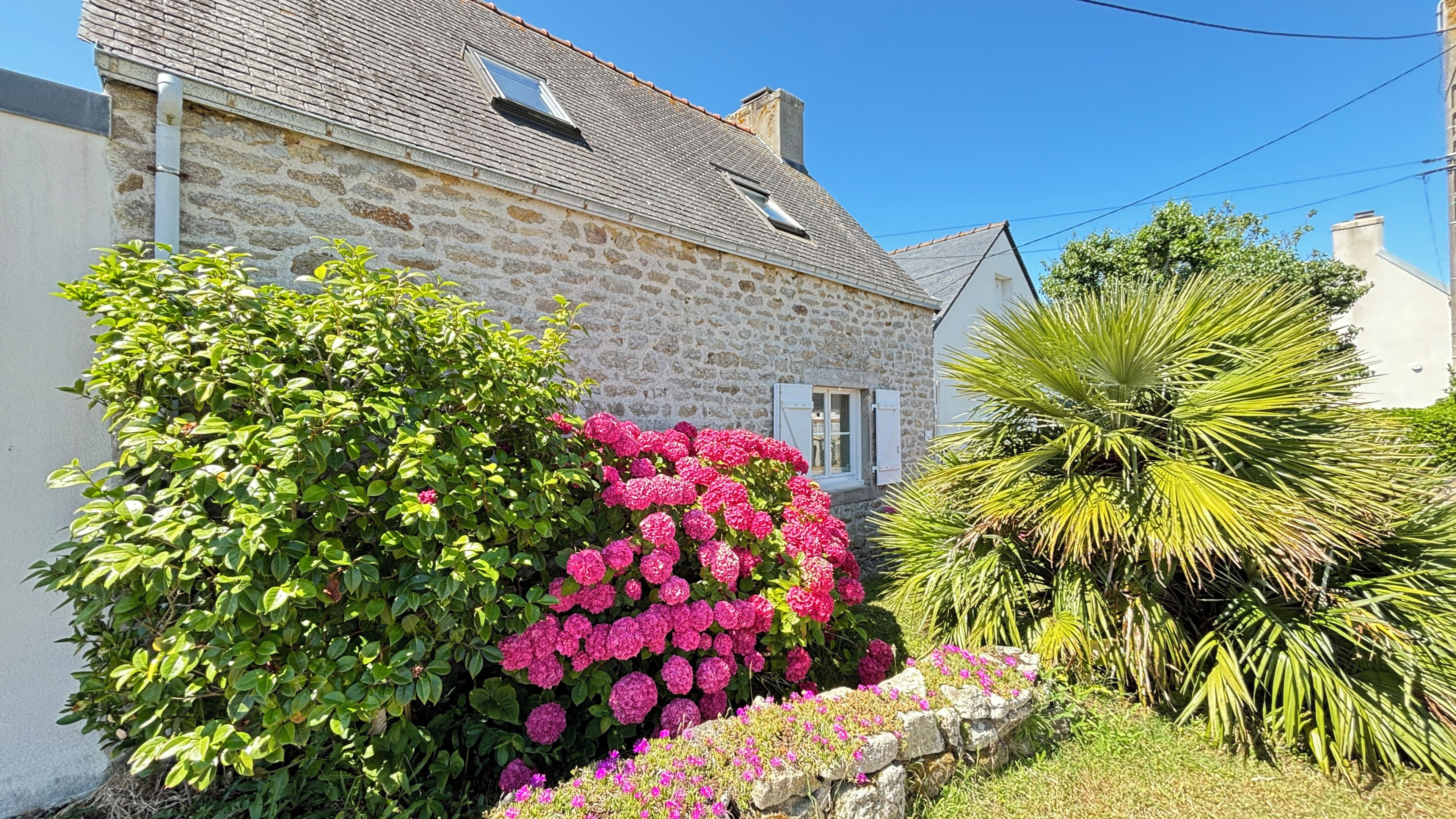 A stone house with a sloped roof, a small window, and a garden with various plants and flowers.