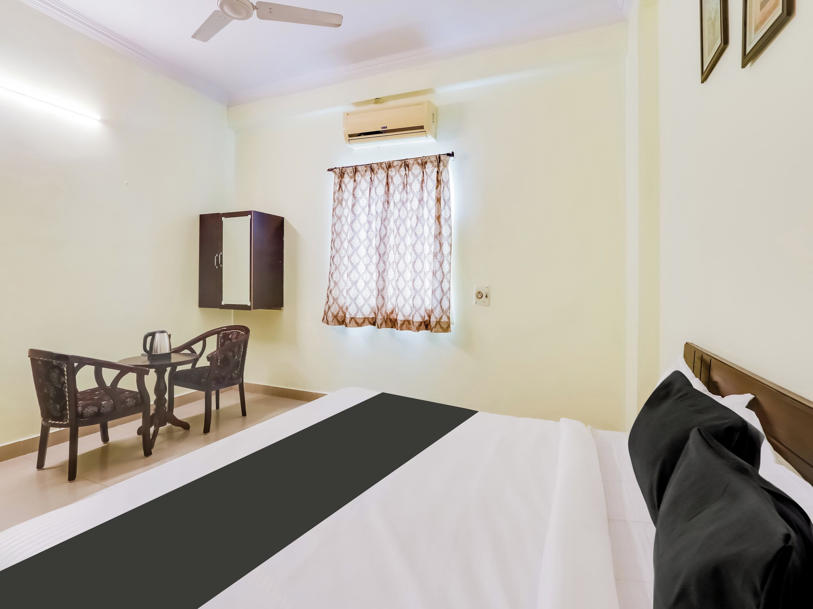 A hotel room with a bed, a desk with a chair, an air conditioning unit, and a window with patterned curtains.
