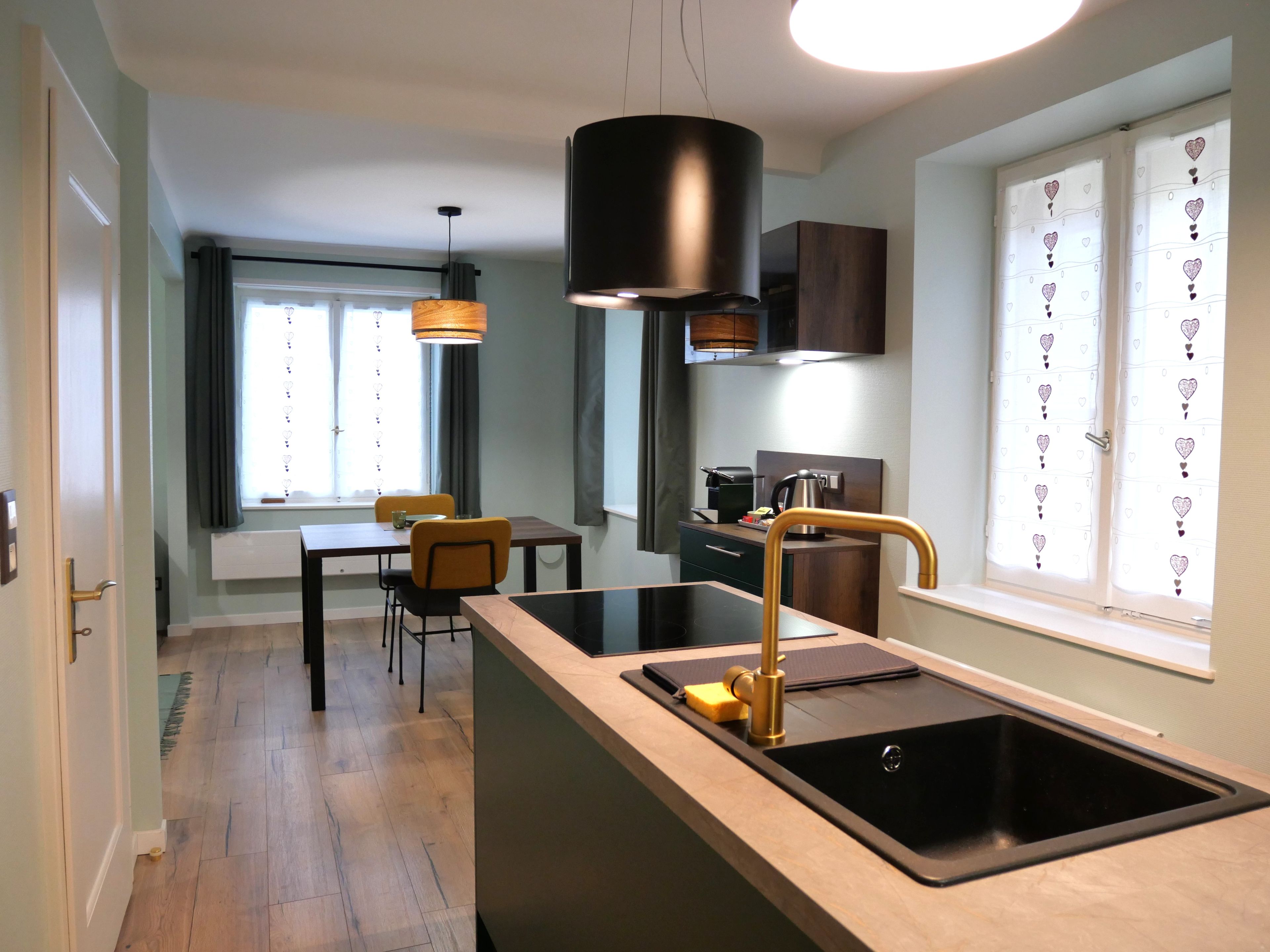 Une cuisine moderne dotée d’un îlot central, d’un évier noir et d’un robinet doré. On y trouve également un coin repas avec une table et des chaises, ainsi qu’une fenêtre agrémentée de rideaux.