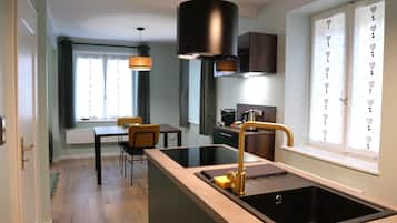 A modern kitchen with a central island, a black sink, and a gold faucet. There is a dining area with a table and chairs, and a window with curtains.
