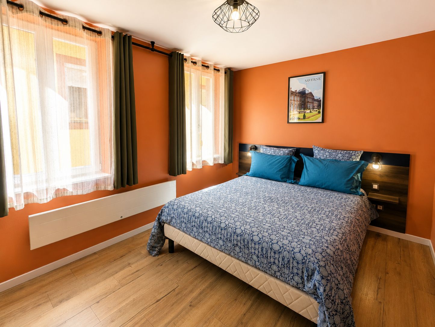 Une chambre à coucher comprenant un lit, des rideaux, un radiateur, une tête de lit, une table de chevet et un luminaire fixé au mur.