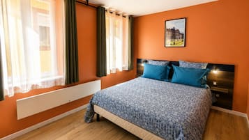 A bedroom with a bed, curtains, a radiator, a headboard, a nightstand, and a wall-mounted light fixture.