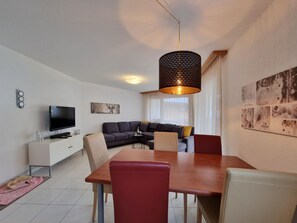 Ein modernes Wohnzimmer mit Flachbildfernseher, einer Couch, einem Esstisch und einer Pendelleuchte.