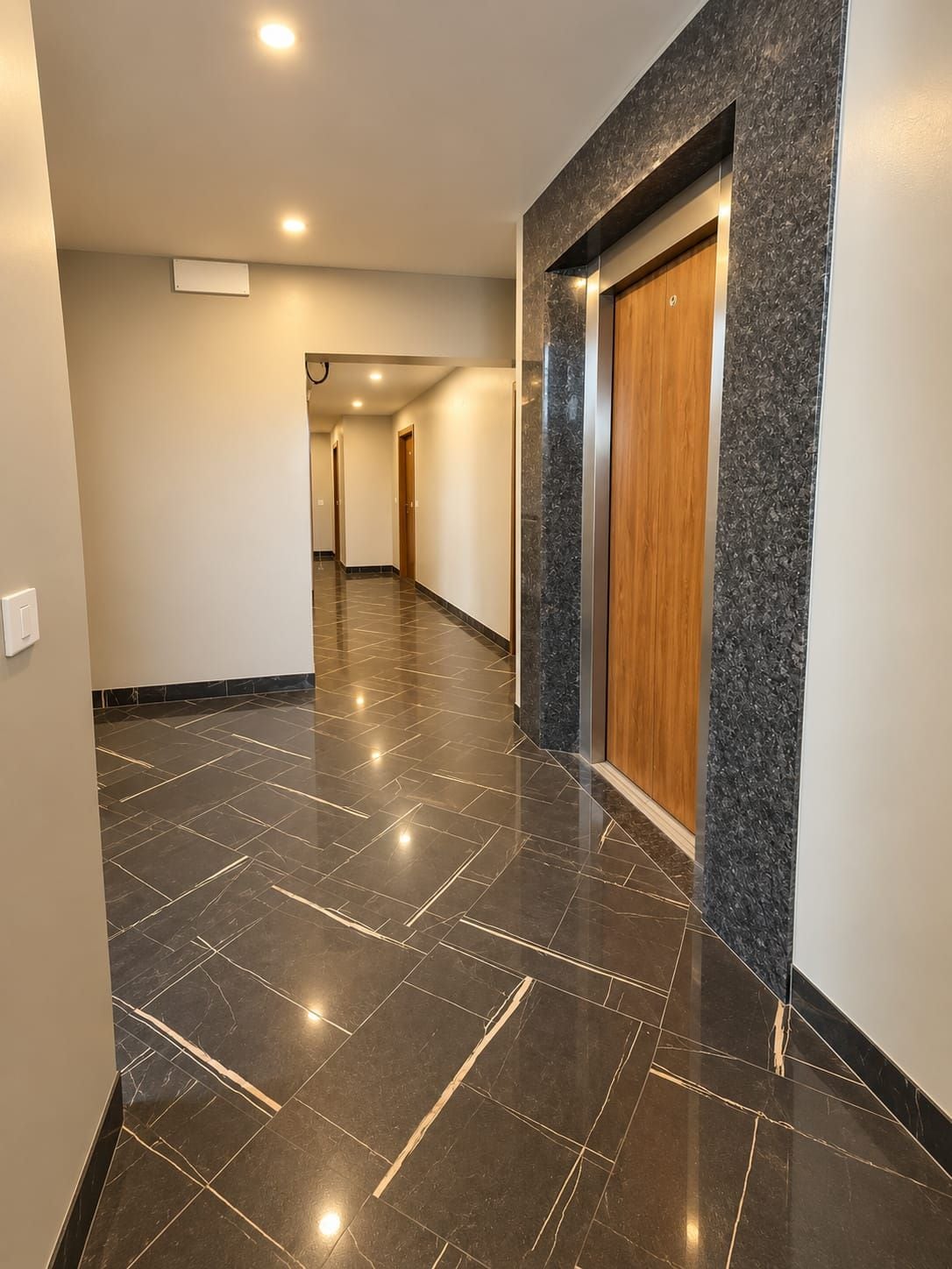 Un couloir doté d’un sol carrelé noir poli, d’une porte en bois et d’un mur décoratif en marbre.
