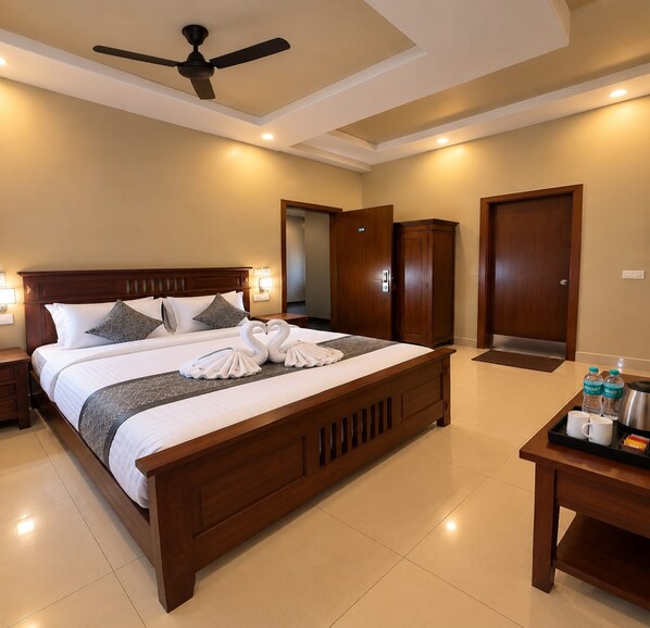 A hotel room with a large bed, a ceiling fan, a nightstand, a bedside table with a tray of amenities, and a wooden wardrobe. - Hotel Grand Mirage  (Chennai)
