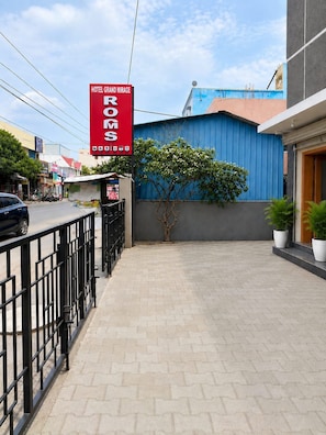 A hotel entrance with a red sign, a paved walkway, a metal fence, and a blue building in the background. - Hotel Grand Mirage  (Chennai)