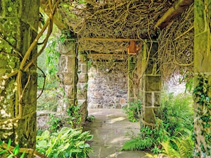 A stone pathway leading through a lush garden with dense foliage and a wooden structure overhead. - Baillie Scott Cottage - UKC10041 (St Andrews)