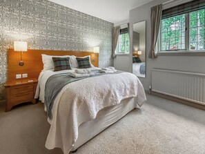 A neatly made bed with a patterned bedspread and pillows, a bedside lamp, a nightstand, and a window with curtains. - Baillie Scott Cottage - UKC10041 (St Andrews)