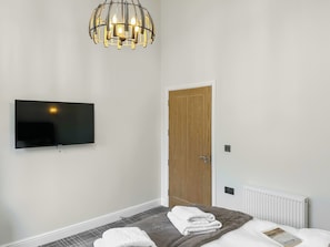 A hotel room with a flat-screen TV mounted on the wall, a wooden door, a radiator, and a bed with a grey blanket and white towels. - 3 bedroom accommodation in Eyemouth (Eyemouth)