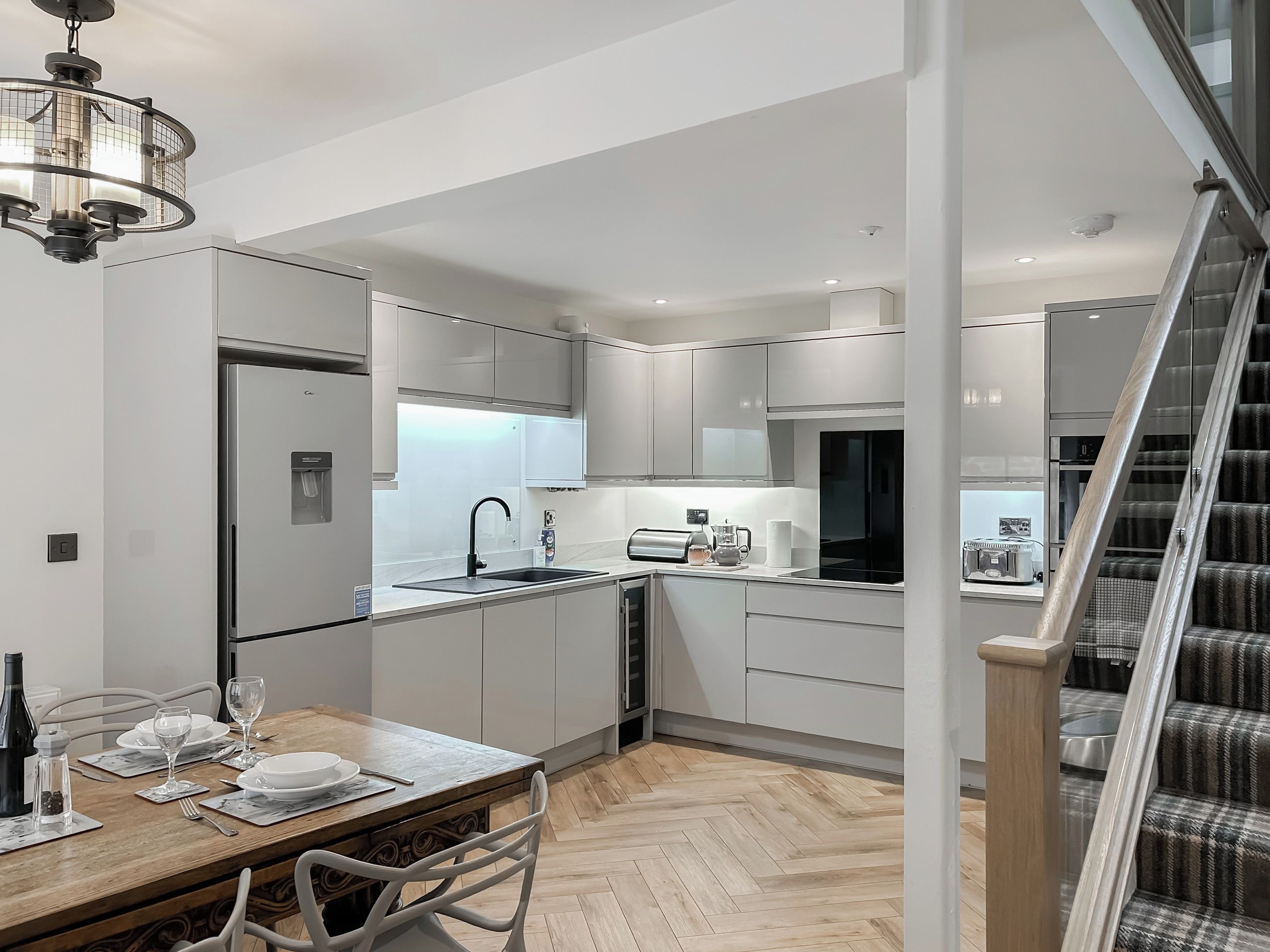 A modern kitchen with white cabinetry, a wooden dining table, and a staircase leading to another level.