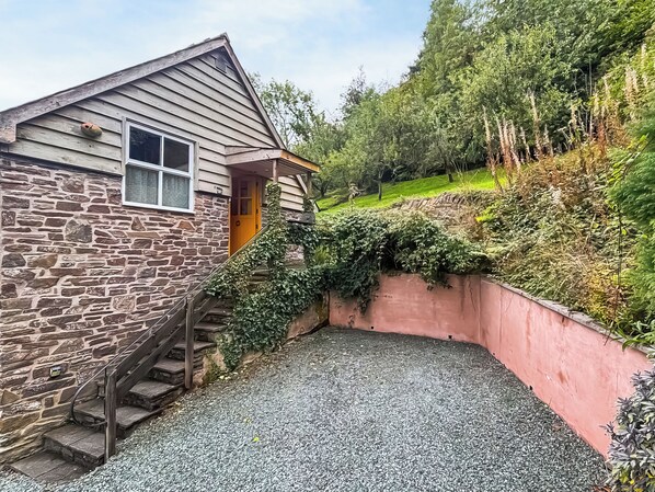 A stone building with a wooden staircase leading to a door, surrounded by greenery and a gravel driveway. - 1 bedroom accommodation in Bishops Castle (Bishops Castle)