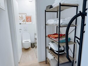 A bathroom with a toilet, a shelf with folded towels, and a vacuum cleaner. - Spacious modern 3-bedroom apartment in beautiful Zadar with AC in every room! (Zadar)