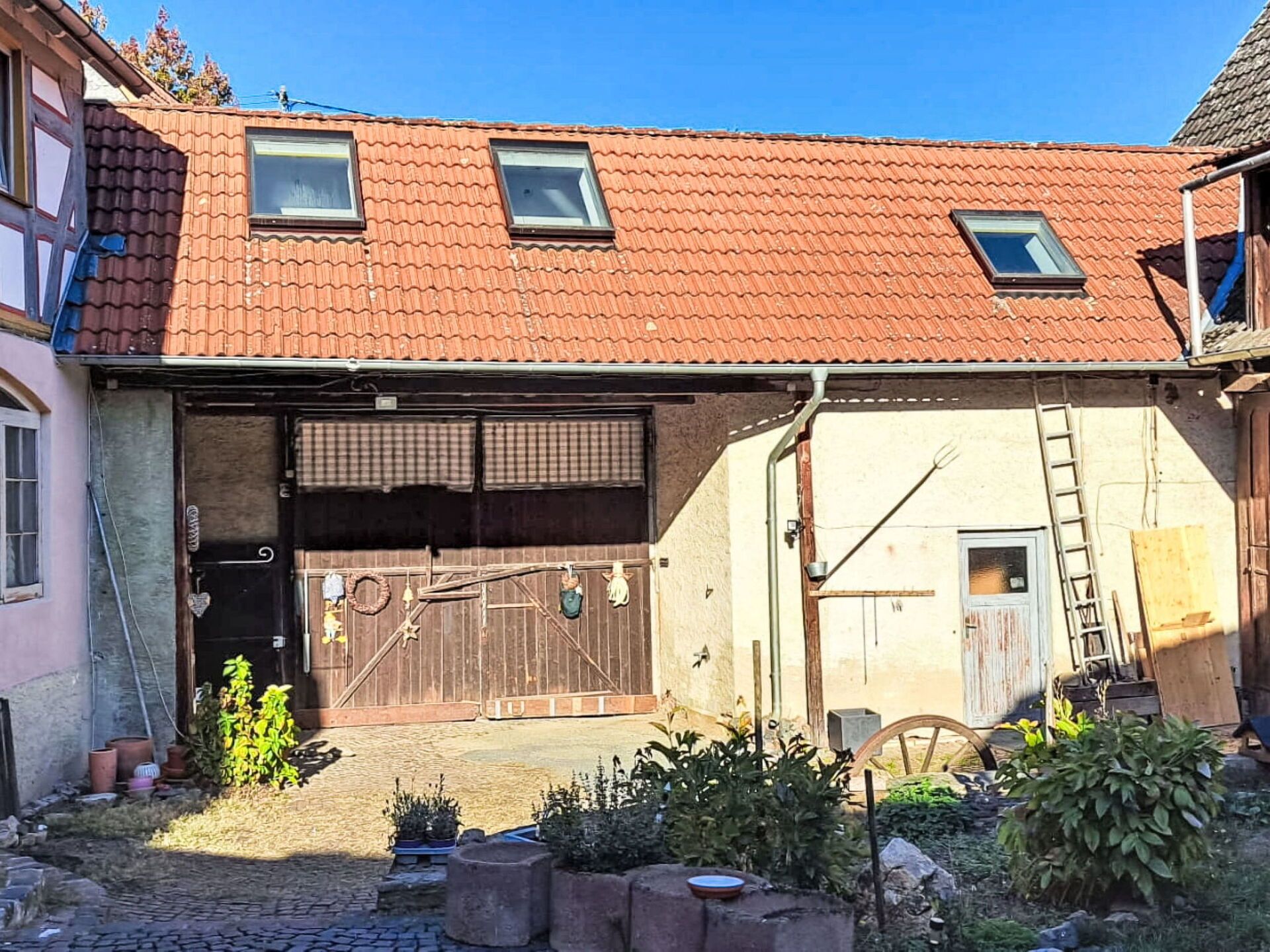 Un bâtiment à deux étages, avec un toit recouvert de tuiles rouges, un garage dont le volet est fermé, et une échelle appuyée contre le mur.