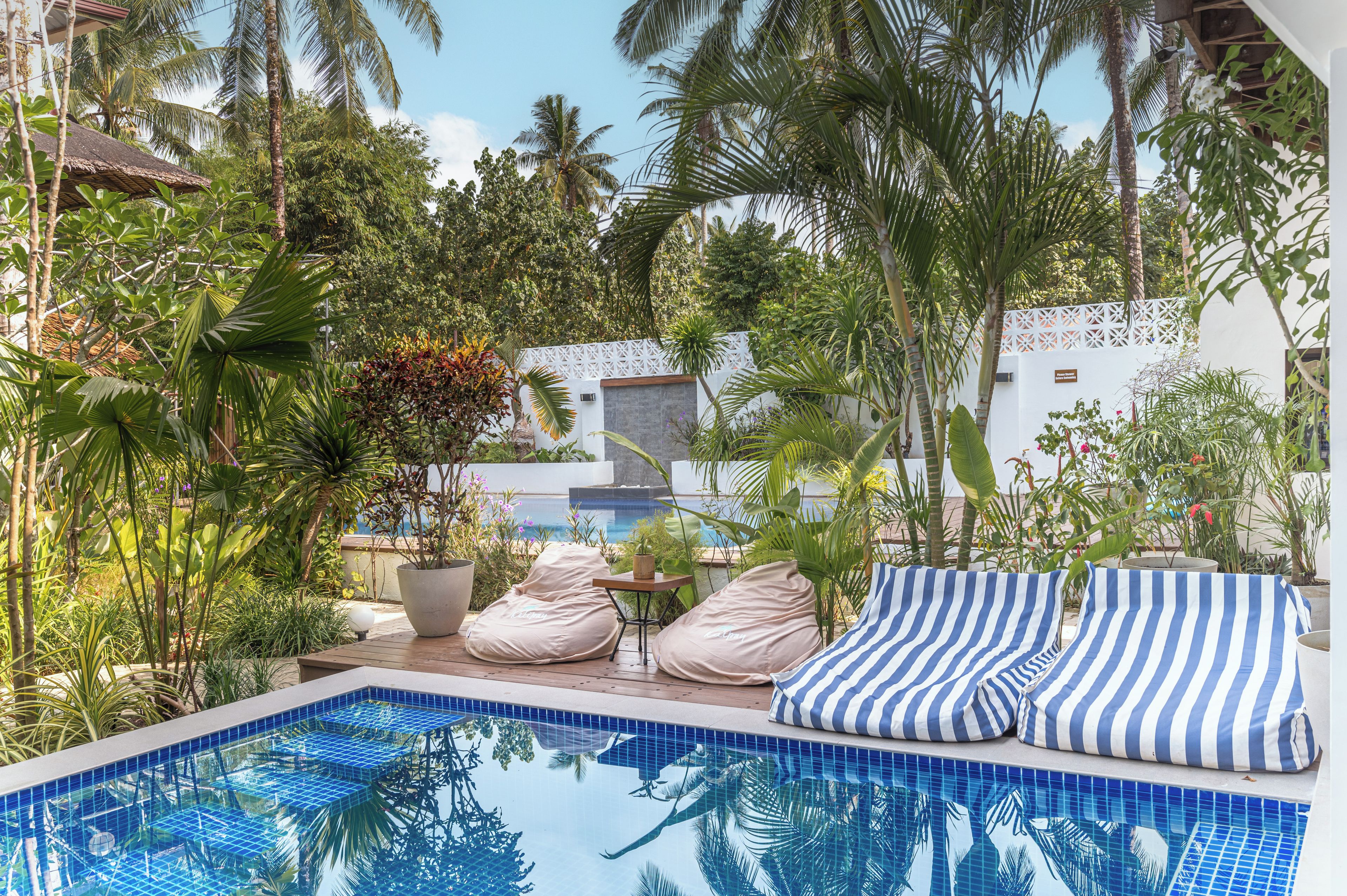 Un área de piscina con sillones reclinables y una mesita, rodeada de plantas tropicales y una valla blanca.