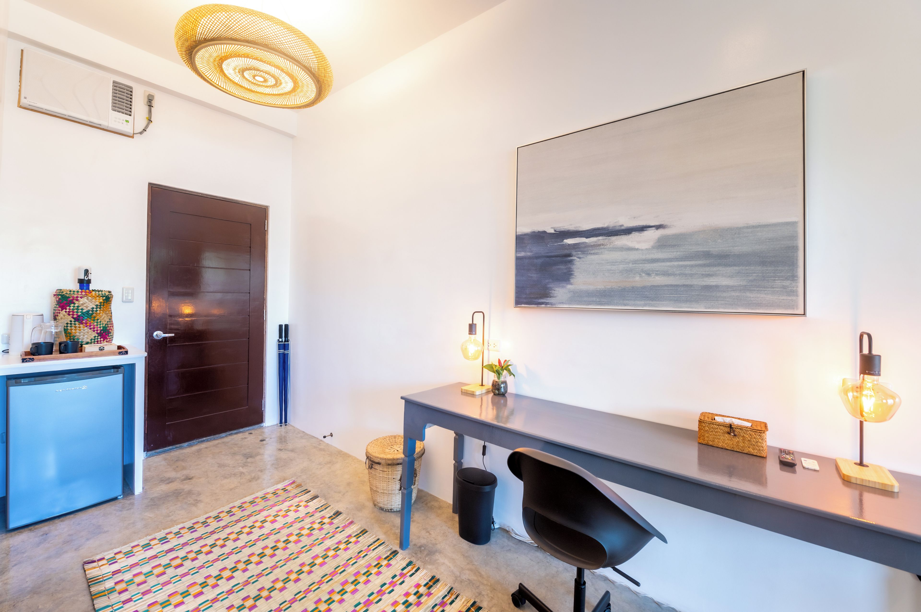 Een moderne keuken met een ingebouwde air conditioner, een donker houten deur, een kleurrijke geweven mand, een kleine koelkast en een vloerkleed met dessin.