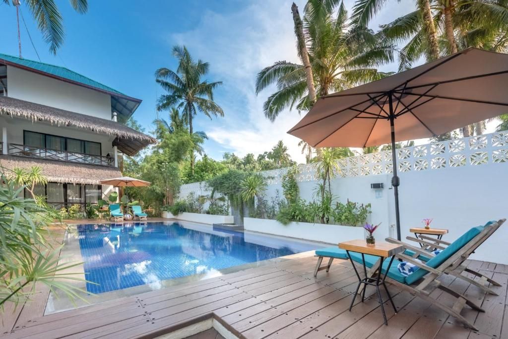 A swimming pool with lounge chairs and umbrellas, surrounded by palm trees and a white fence.