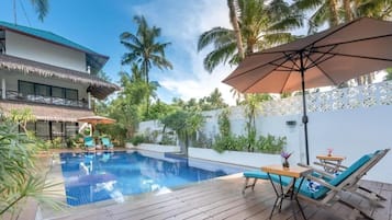 A swimming pool with lounge chairs and umbrellas, surrounded by palm trees and a white fence.