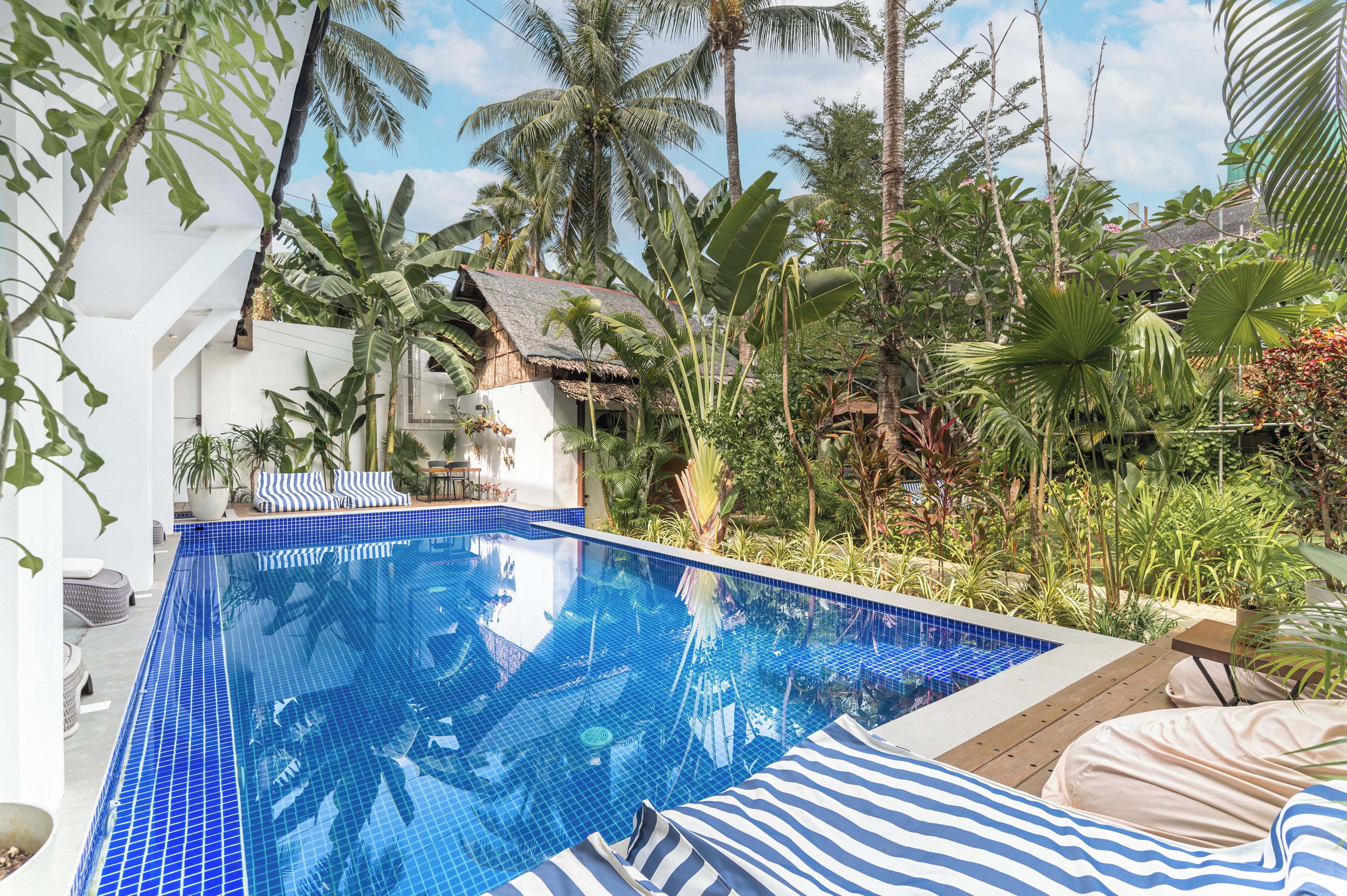 Una piscina con azulejos azules, rodeada de plantas tropicales y un edificio blanco con techo de paja.