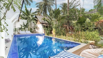 Una piscina con azulejos azules, rodeada de plantas tropicales y un edificio blanco con techo de paja.