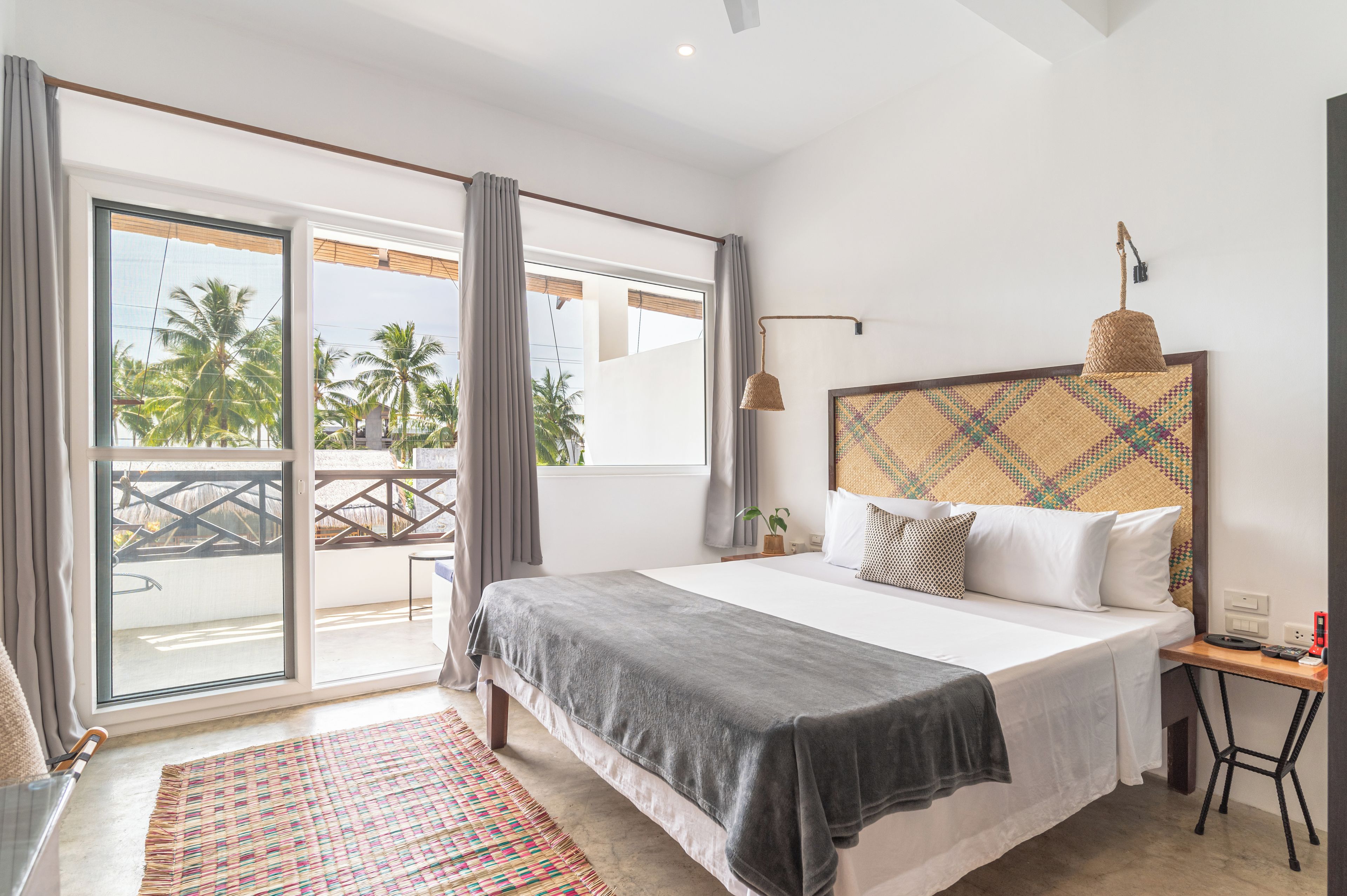 A bedroom with a bed, a bedside table, a lamp, a window with curtains, and a view of palm trees.