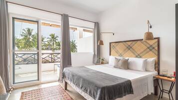 A bedroom with a bed, a bedside table, a lamp, a window with curtains, and a view of palm trees.